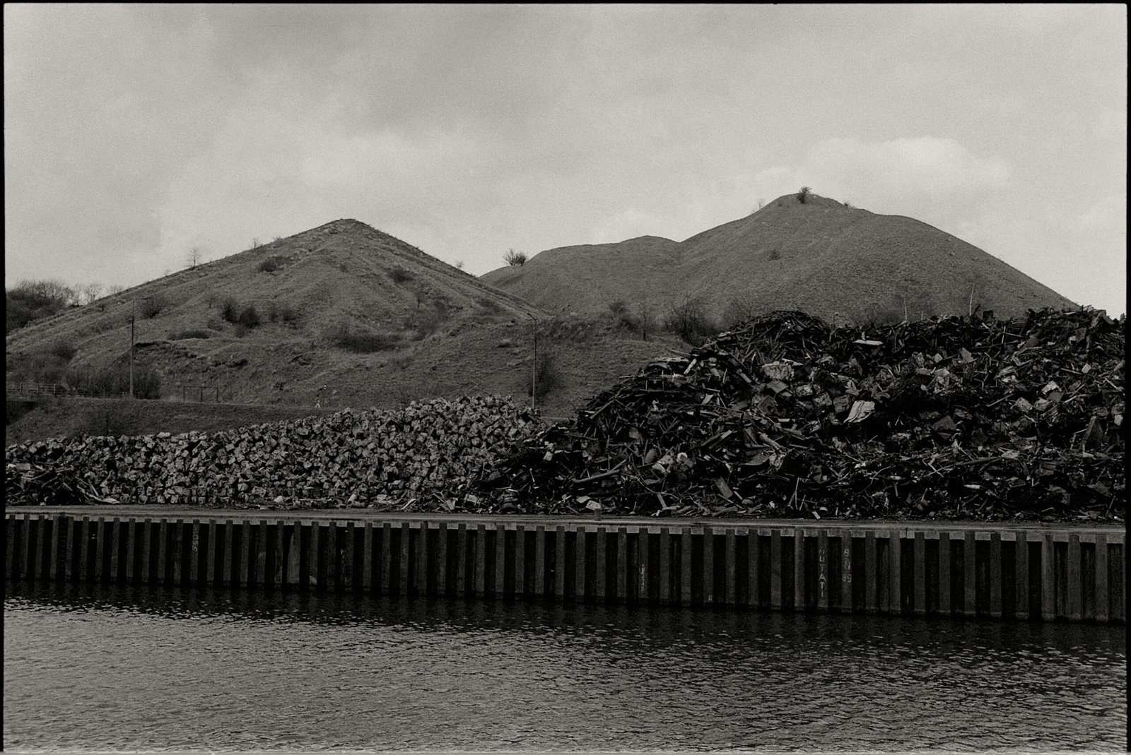 A black and white scene showing hills by a canal with metal scrap heaps on the riverbank.